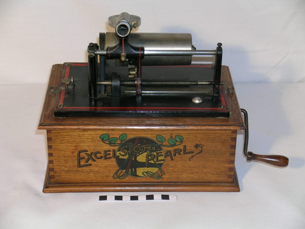 Excelsiorwerke phonograph. circa 1904 A small phonograph with the motor concealed in a wooden case. Black cast-iron bedplate with a red lining. Aluminium mandrel. 'Excelsior Pearl' written in gold lettering overlays a landscape / floral motif on the case.