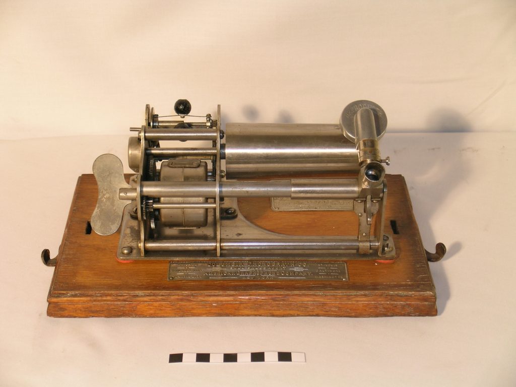 A Columbia Type B Graphophone in a wooden case. The exposed steel plate main frame rests upon a wooden bedplate. Also upon the bedplate is a nickel patent plate and a nickel license plate. Both bedplate and lid are simple, the only decoration being the traditional Graphophone decal garland.